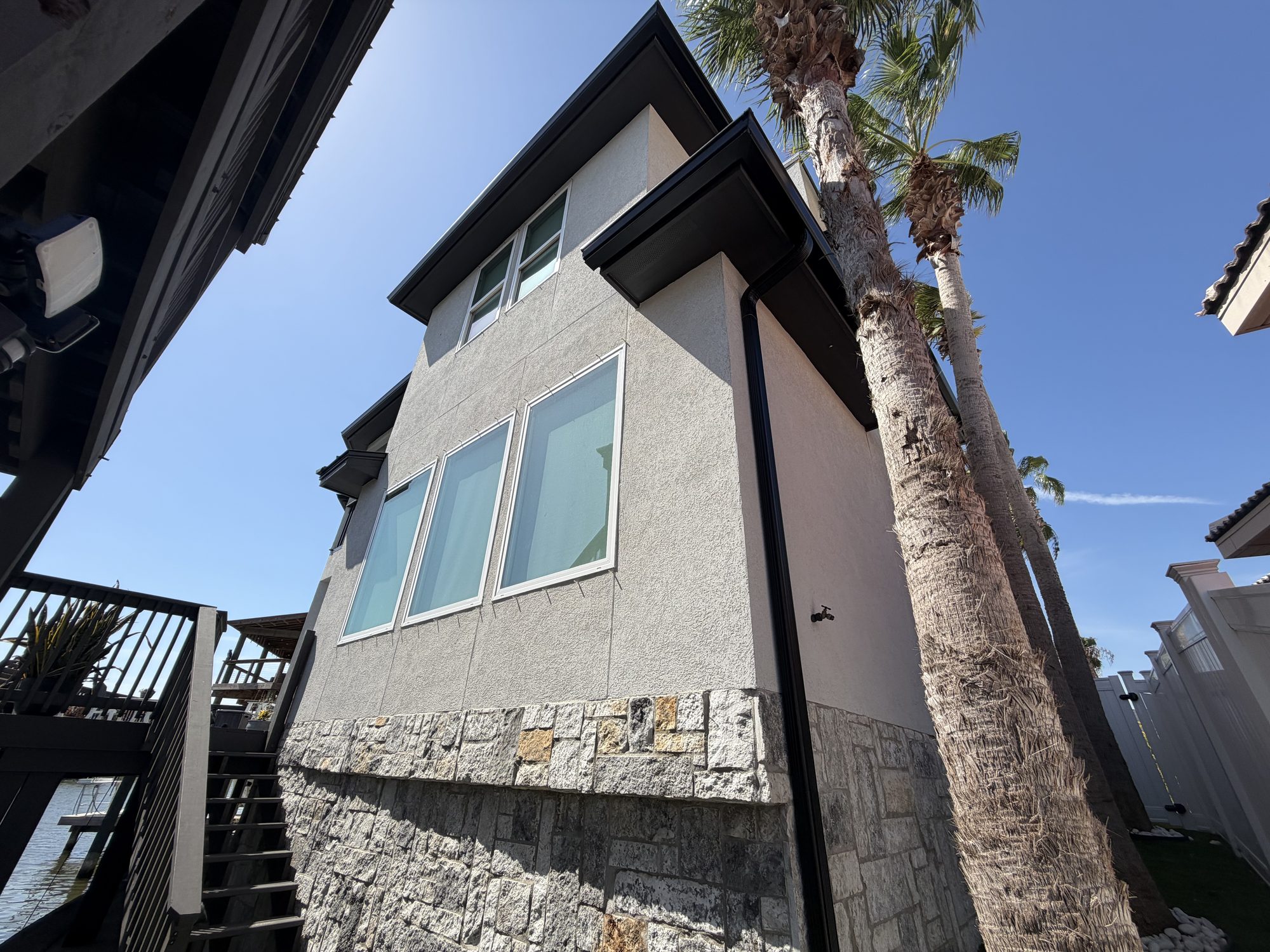 Tall black downspout on a two-story stucco and stone wall with a palm